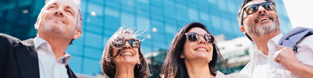 Four adults in business attire and sunglasses stand outdoors, smiling, with a modern glass building in the background.