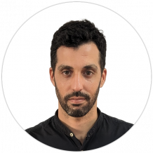 A man with short dark hair, a beard, and mustache is wearing a black buttoned shirt, looking directly at the camera against a plain white background.