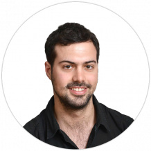 A man with short dark hair and a beard, wearing a black shirt, is smiling at the camera against a plain white background.